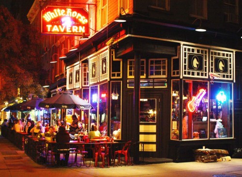 A lively tavern at night, with neon signs and outdoor seating filled with patrons enjoying the atmosphere.