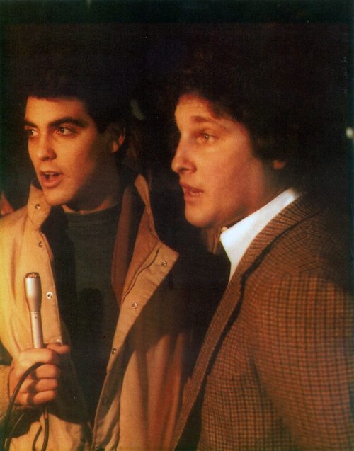 Two young men speaking, one holding a microphone, both dressed in stylish 1970s attire.
