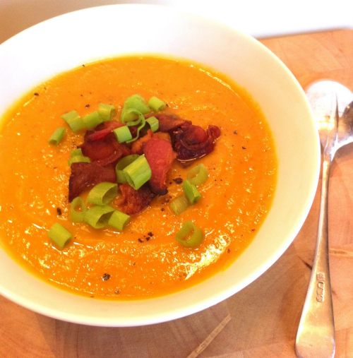 A bowl of creamy orange soup topped with crispy bacon and green onions, served with a silver spoon on the side.