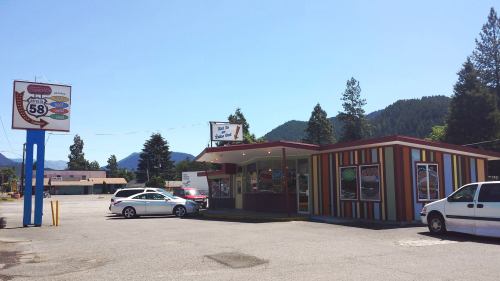 A colorful fast-food restaurant with a sign, surrounded by trees and mountains under a clear blue sky.