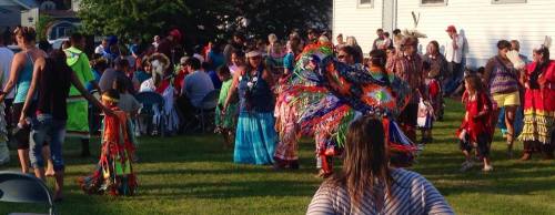 A vibrant outdoor gathering with people in traditional attire dancing, surrounded by a crowd and green grass.