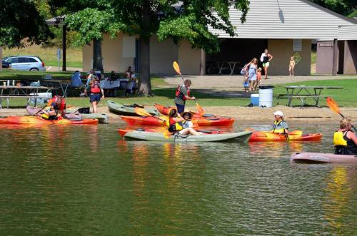 A group of people kayaking on a calm lake, with a park and picnic area in the background.