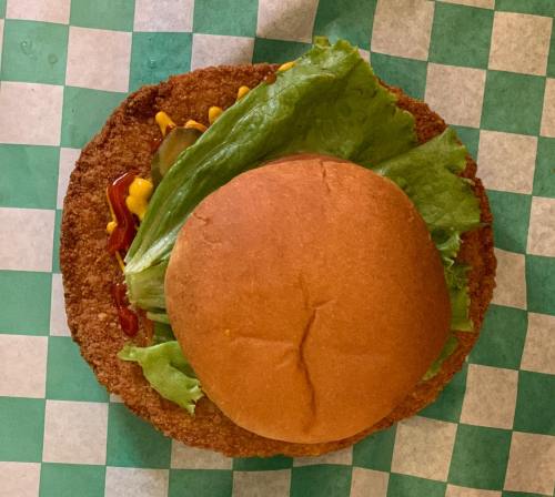 A sandwich with a round bun, lettuce, ketchup, and mustard on a crispy, golden-brown base on a checkered green background.