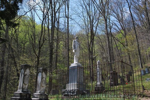 A statue surrounded by gravestones in a wooded cemetery, with trees and a clear blue sky in the background.