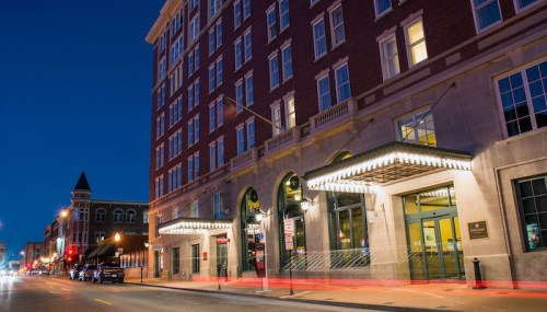 A historic building at night, featuring bright lights and a welcoming entrance, with city streets and traffic nearby.