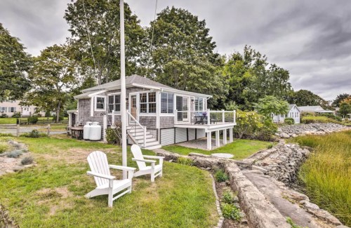 A cozy waterfront cottage with a deck, surrounded by greenery and two white Adirondack chairs in the foreground.