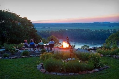 A group of four people sitting by a fire pit, overlooking a river and sunset, surrounded by lush greenery.