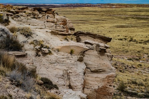 A rocky cliff edge overlooks a vast, grassy landscape under a blue sky.