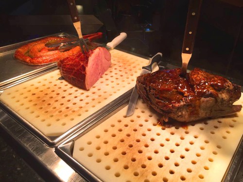A selection of roasted meats on cutting boards, including a large roast, ham, and sausage, with carving knives.