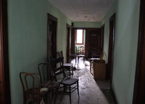 An empty, dimly lit hallway with scattered chairs and a box, featuring peeling green walls and closed doors.