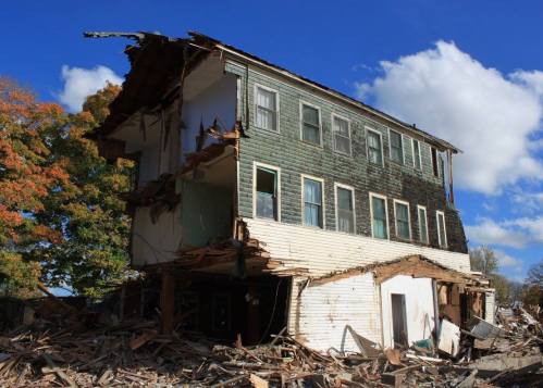 A partially collapsed building with exposed interior, surrounded by debris under a blue sky with scattered clouds.