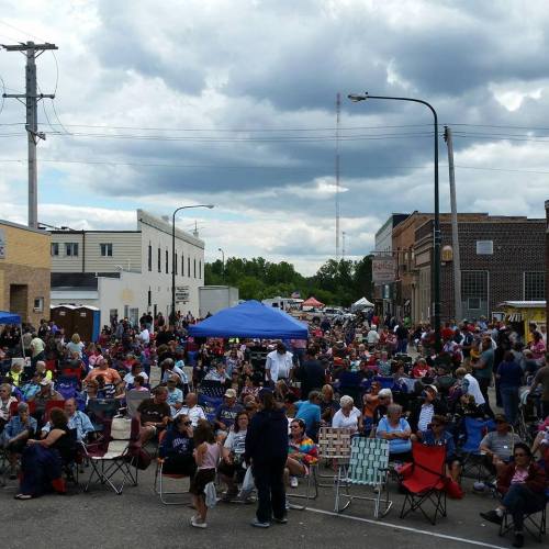 A large crowd gathers in a town square under cloudy skies, with tents and buildings in the background.