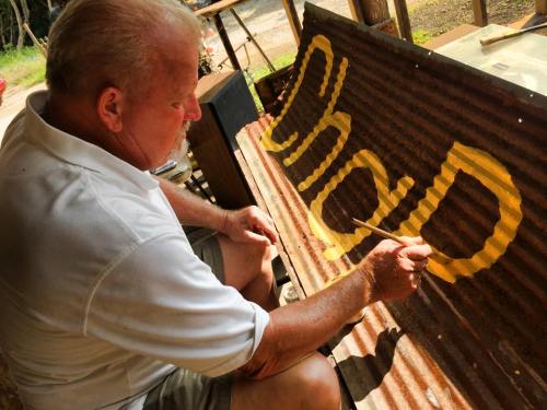 A man paints the word "shed" in yellow on a corrugated metal surface outdoors.