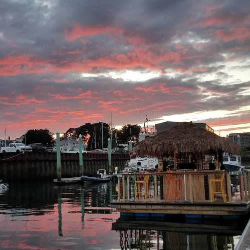 A vibrant sunset over a marina, reflecting on calm waters, with a thatched-roof structure in the foreground.