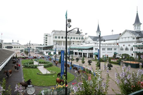A view of a horse racing venue with horses, spectators, and lush gardens under a cloudy sky.