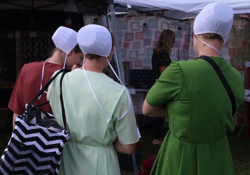 Three women in traditional dresses and bonnets stand together, facing away, at a market under a tent.