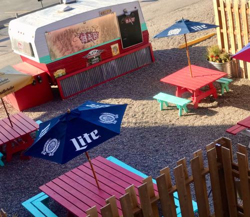 Colorful outdoor seating area with red and turquoise tables, umbrellas, and a food truck in a gravel setting.