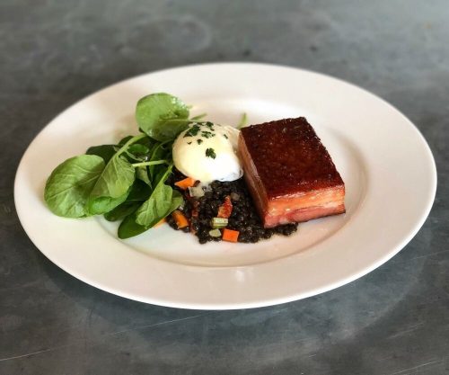 A plate featuring crispy pork belly, poached egg, lentils with vegetables, and fresh greens.