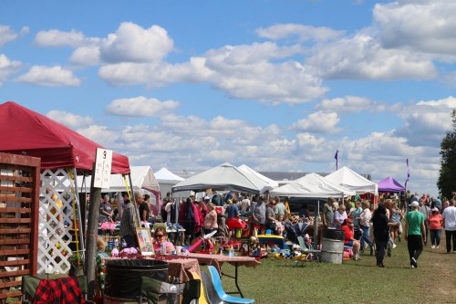 A bustling outdoor market with colorful tents, vendors, and crowds under a blue sky with fluffy clouds.