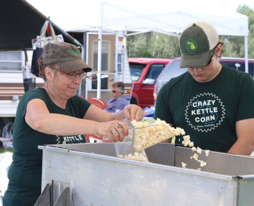 A woman and a man in matching green shirts scoop popcorn from a kettle at a fair or market.
