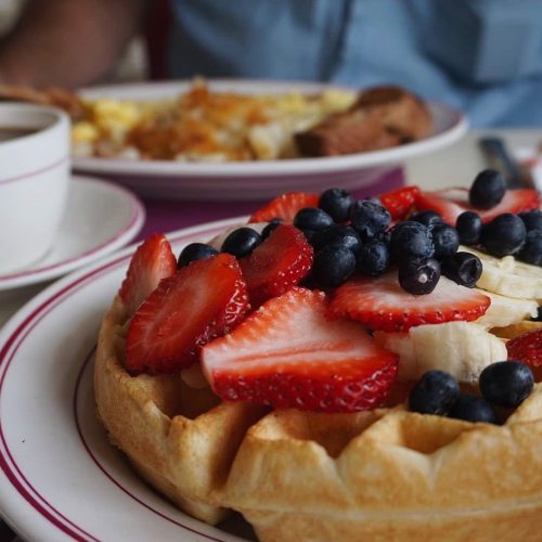 A close-up of a waffle topped with strawberries, blueberries, and banana slices, with a coffee cup in the background.