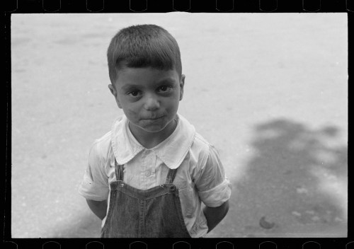 A young boy with short hair stands outdoors, wearing a white shirt and denim overalls, hands behind his back.