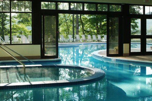 Indoor pool area with a hot tub, large windows, and a view of an outdoor pool surrounded by lounge chairs.