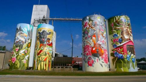 Colorful murals on silos depict animals and nature, set against a blue sky and industrial backdrop.