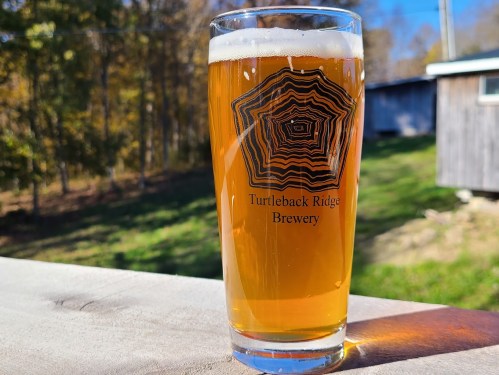 A glass of beer from Turtleback Ridge Brewery sits on a wooden surface, with a blurred outdoor background.