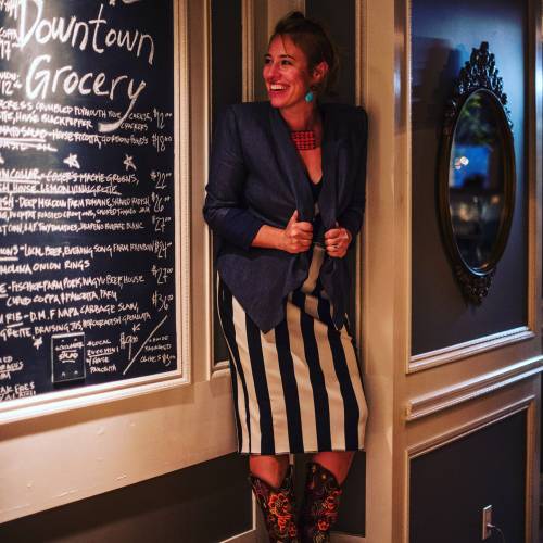 A woman in a blue blazer and striped skirt smiles playfully next to a chalkboard menu in a stylish setting.