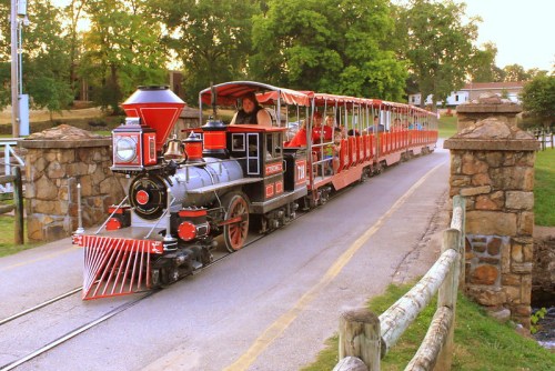 A vintage steam train with a red and gray engine crosses a stone bridge, surrounded by greenery and a scenic landscape.
