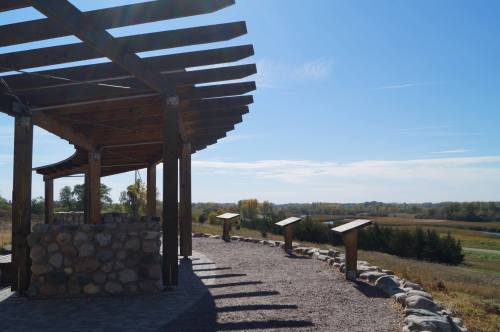 A scenic view from a wooden pavilion overlooking a grassy landscape and river under a clear blue sky.