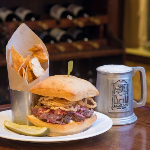 A hearty sandwich with sliced beef, crispy onion rings, a side of potato chips, and a frosty mug of beer.
