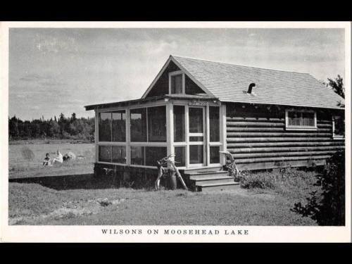 Black and white photo of a log cabin by Moosehead Lake, with a porch and a view of the water in the background.