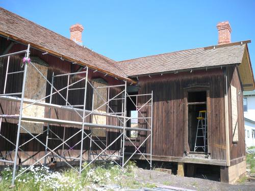 A wooden building under renovation, featuring scaffolding, boarded windows, and a ladder against the wall.