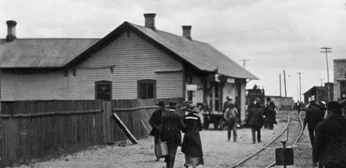 Black and white photo of people walking along a railway track near a station, with buildings in the background.