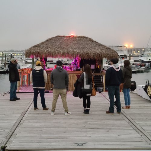 A group of people stands on a dock near a thatched-roof bar, with boats and a sunset in the background.