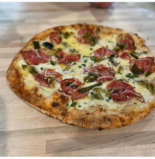 A freshly baked pizza topped with pepperoni, green peppers, onions, and herbs on a wooden table.
