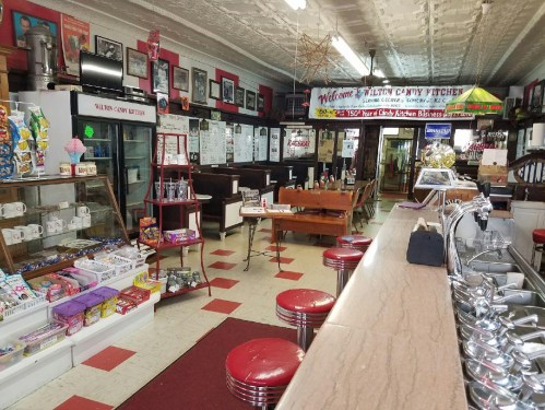 A vintage candy shop interior with a counter, red stools, shelves of sweets, and nostalgic decor.