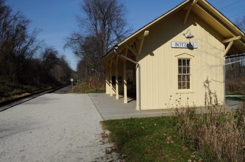 A yellow train station building beside a gravel path, with a railway track visible in the background and trees nearby.