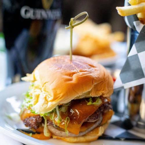A close-up of a juicy cheeseburger with lettuce and sauce, garnished with a toothpick, served with fries and a pint of Guinness.