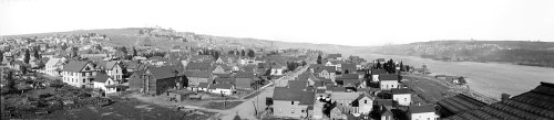A panoramic view of a small town with houses lining a riverbank, surrounded by hills in the background.