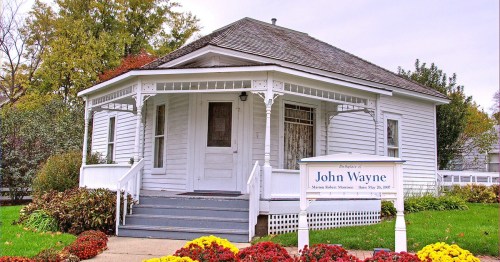 Historic white house with a porch, labeled as the birthplace of John Wayne, surrounded by colorful flowers.
