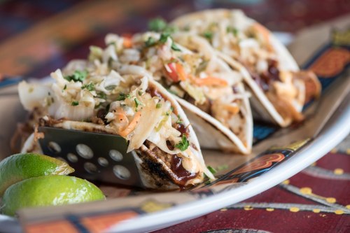 Three tacos filled with shredded cabbage and colorful toppings, served with lime wedges on a decorative plate.