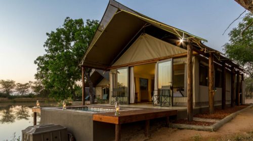 A modern safari lodge by a tranquil lake, featuring large windows and a wooden deck with chairs.