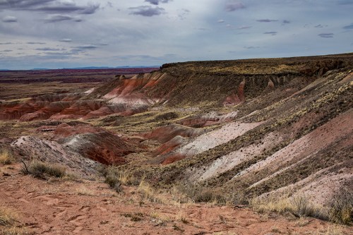 A rugged landscape featuring colorful, layered hills and valleys under a cloudy sky. Sparse vegetation is visible.