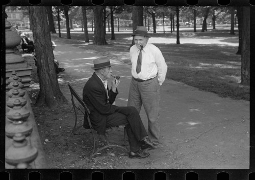Two men in hats converse in a park, one seated on a bench smoking a pipe, the other standing nearby.