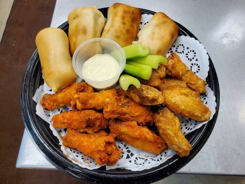 A platter of crispy chicken wings, celery sticks, a cup of ranch dressing, and soft bread rolls on a decorative tray.