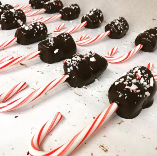 Chocolate-covered treats on candy cane sticks, topped with crushed peppermint, arranged on a white surface.