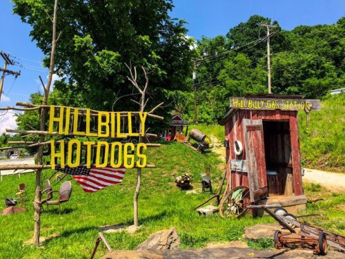 A colorful roadside stand with a sign reading "Hillbilly Hotdogs" and a rustic shed, surrounded by greenery.